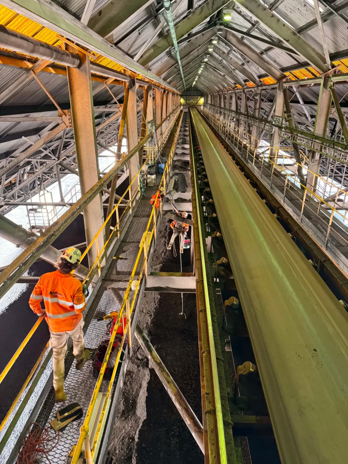 Konstruct technicians working along the walkway of an isolated coal bunker conveyor during a shutdown, one crew member suspended on rope beside the belt