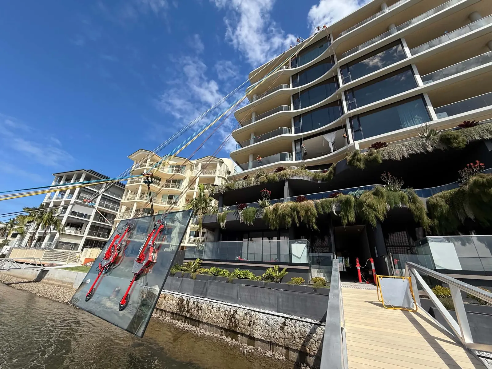 Two red vacuum-lifter rigs lowering a 900-kilogram glass unit over the water beside a Maroochydore canal-front building